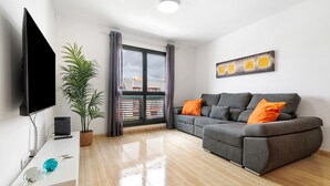 A modern living room with a flat-screen TV, grey sofa with orange cushions, a potted plant, and a large window with curtains.