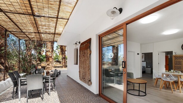 An open-plan living area with a dining set, a glass door leading to a kitchen, and a patio with outdoor furniture.