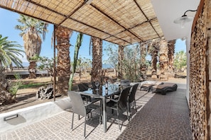 Un patio exterior con mesa y sillas de vidrio, rodeado de palmeras y vista al mar.