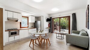 A modern kitchen with white cabinets, a dining table with chairs, a grey sofa, and a TV mounted on the wall.