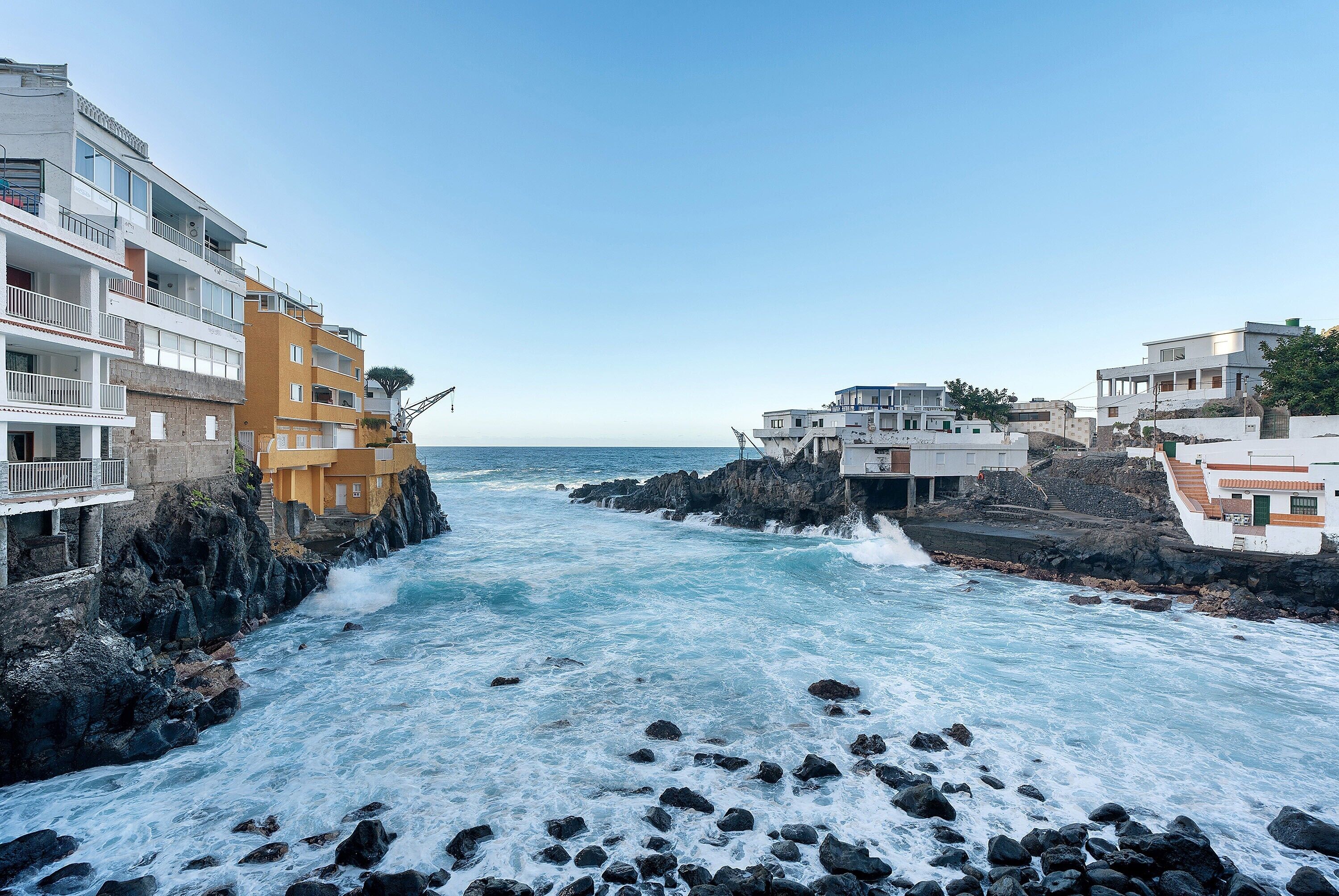 A coastal town with buildings along the rocky shoreline, a clear sky, and waves crashing against the rocks.