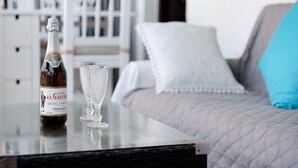 Una botella de Sidra El Gaiter sobre una mesa con un vaso transparente a su lado, en una habitación con cama y muebles blancos.