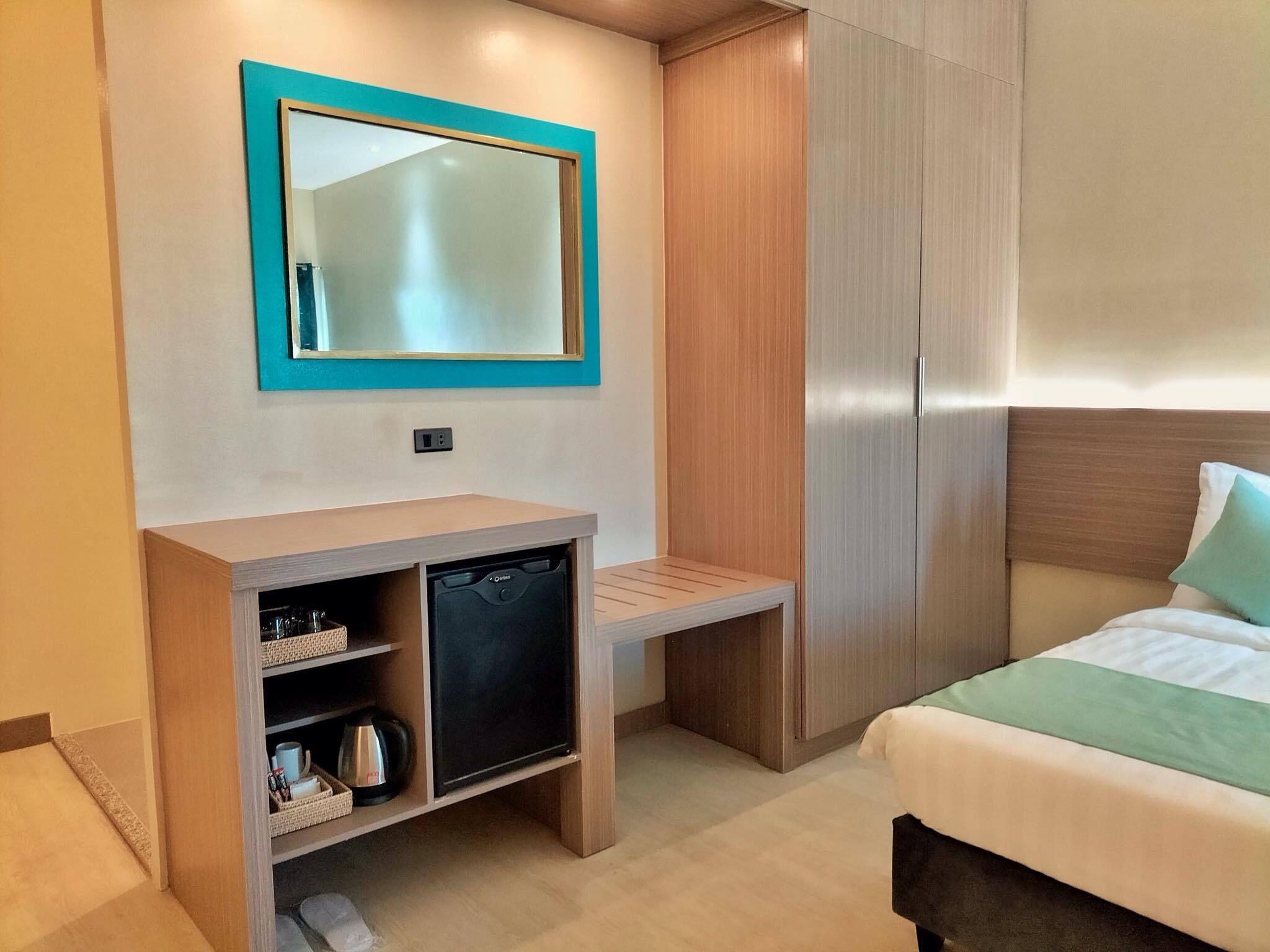 A compact hotel room with a bed, a small desk with a dishwasher, a mirror, and a wardrobe.