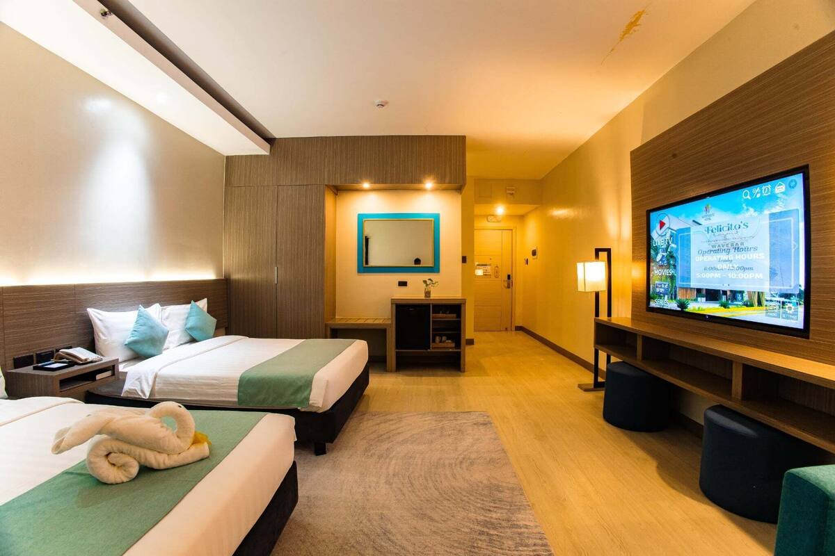 Une chambre d’hôtel avec deux lits, une télévision, un miroir, une petite table et un écran fixé au mur qui affiche des informations sur l’hôtel.
