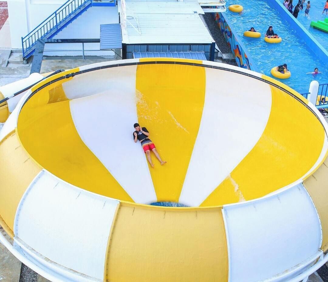 Un grande scivolo acquatico con sezioni gialle e bianche, una persona che scende e un'area piscina con persone su gommoni.