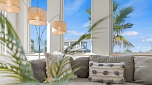 A modern living room with a gray sofa, two decorative pillows, and a large window with a view of palm trees and a house. - Luxury Retreat w/Games, Patio, BBQ, Close to Beach (Palm Coast)