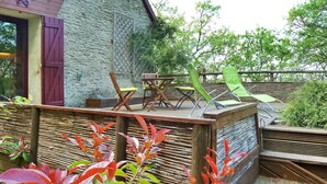 Una terraza de madera con mobiliario exterior, un muro de piedra y abundante vegetación.