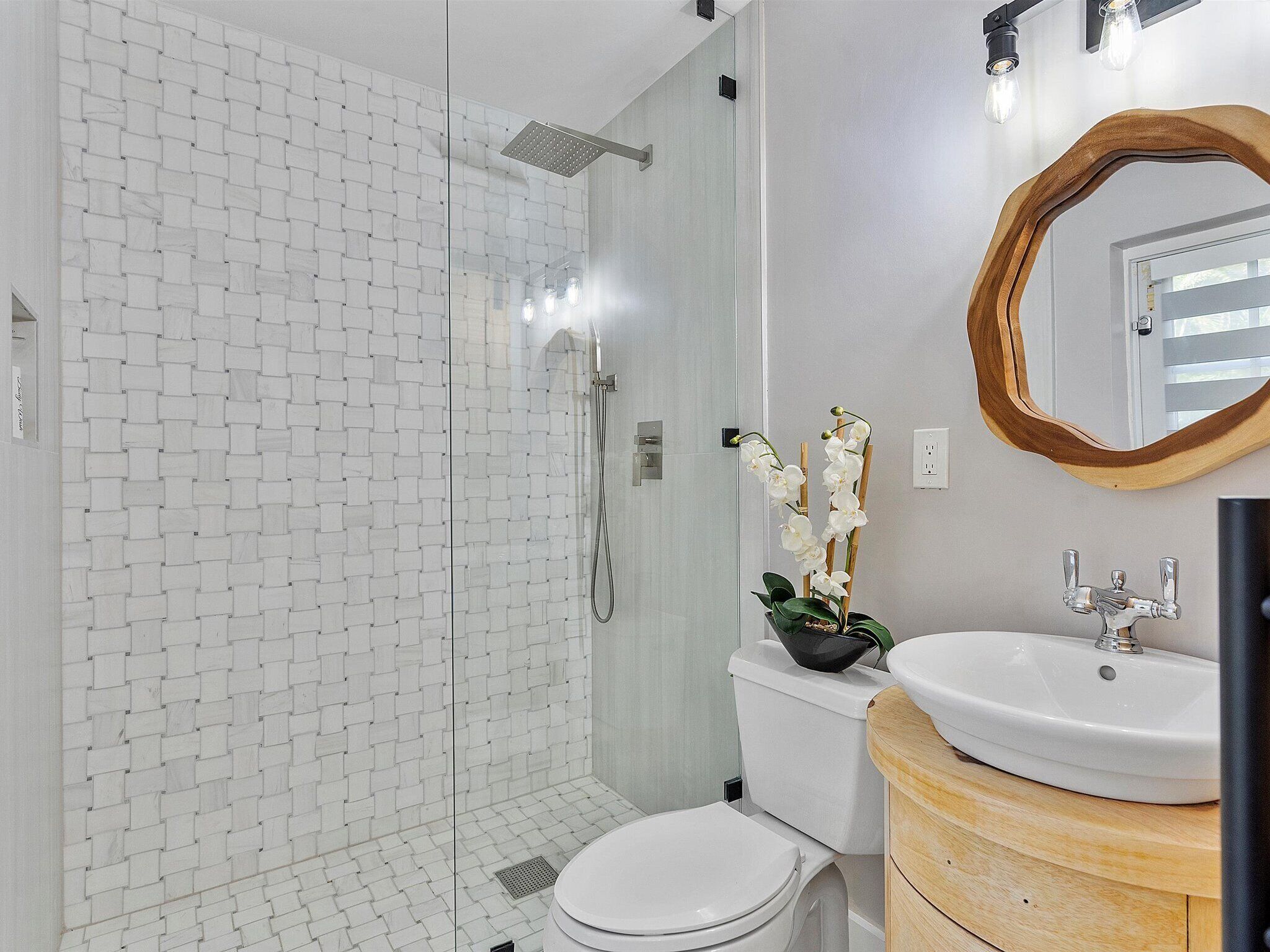 A modern bathroom with a glass shower enclosure, white tiled walls, a white toilet, a wooden vanity with a sink, and a mirror with a wooden frame.