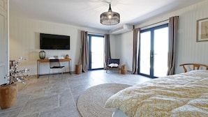 Habitación de hotel con cama, escritorio con silla, televisor, planta en maceta y una ventana grande con cortinas.