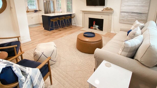 Un salon moderne avec un canapé, un pouf et une table basse. On y trouve également un coin cuisine équipé d’un réfrigérateur, d’un évier et de tabourets de bar. Un téléviseur à écran plat est fixé au mur, et une cheminée est visible.