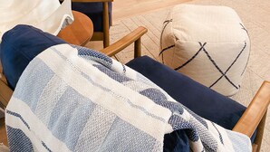 Un salon moderne avec un canapé, une table basse et un plaid à motifs. On aperçoit, au fond, une cuisine avec un coin bar et un réfrigérateur.