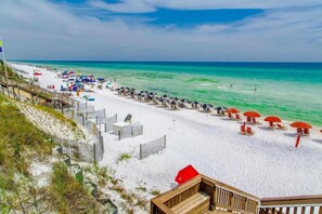Ein Strand mit weißem Sand, klarem türkisblauem Wasser und roten Sonnenschirmen. Holzstege und Liegestühle sind vorhanden.