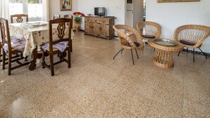 Ein Essbereich mit einem Tisch für vier Personen, umgeben von Korbstühlen mit Kissen. Es steht eine Holzkommode mit einem Fernseher darauf, und an der Wand hängt ein Gemälde.