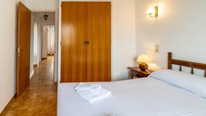 Habitación de hotel con un armario de madera, una cama con ropa blanca, una mesita de noche con una lámpara y un pasillo que conduce a otra habitación.