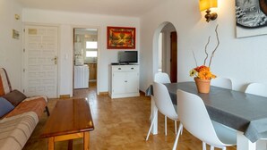 Una sala de estar con un sofá, una mesa de madera y un comedor con sillas blancas y una mesa. Se ve una cocina al fondo con un refrigerador y un televisor.