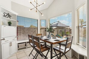 A dining area with a table, chairs, and a chandelier. There are windows with blinds and a view of the outside.