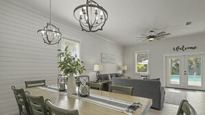 Un espacio habitable amplio con una mesa de comedor, sillas y un sofá. Hay un ventilador de techo, un letrero que dice “Welcome” y una vista de una piscina a través de la puerta de cristal.