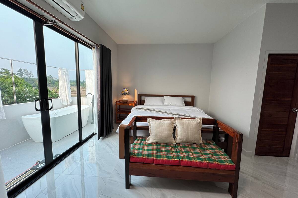 A bedroom with a bed, a bedside table, a lamp, and a view of a balcony with a bathtub.