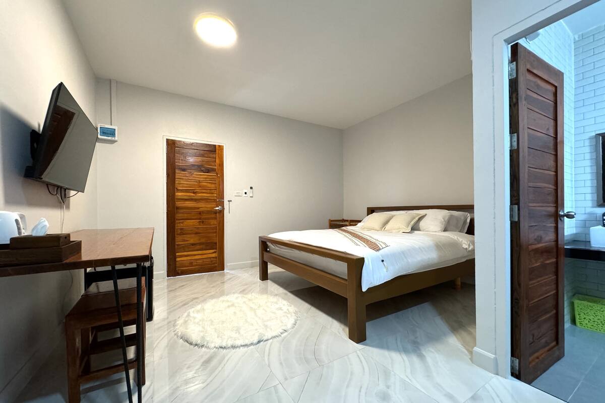 A bedroom with a bed, a desk, a chair, a TV mounted on the wall, and a door leading to another room.