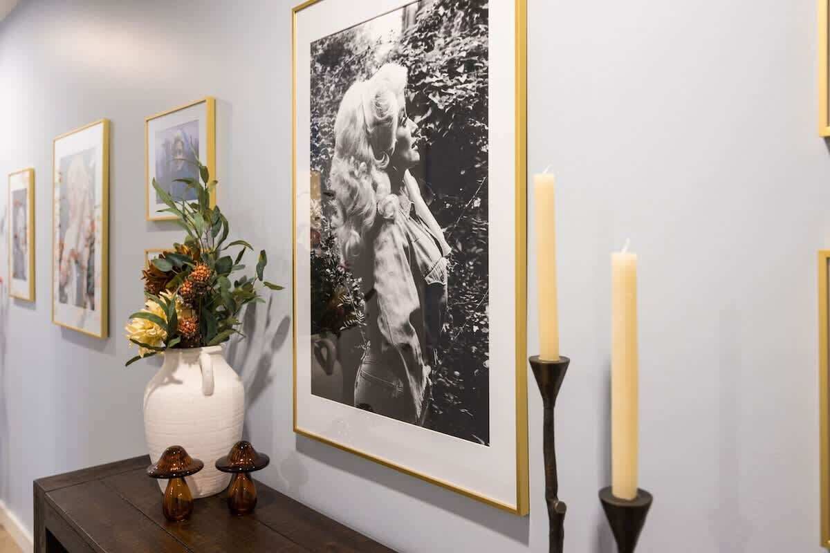 Een gang met ingelijste foto’s aan de muur, een witte vaas met bloemen op een tafel, en twee kaarsen.