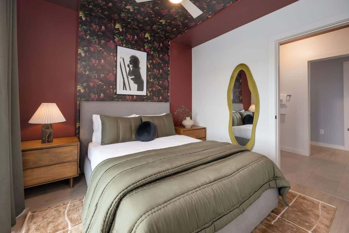 A bedroom with a bed, nightstands, a lamp, a mirror, and a floral-patterned wall.