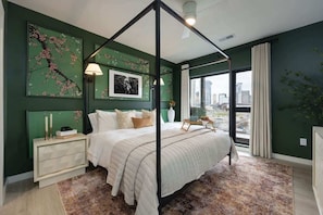 Une chambre à coucher avec un lit à baldaquin, une table de chevet avec une lampe, une grande fenêtre avec des rideaux et des œuvres d’art accrochées aux murs.