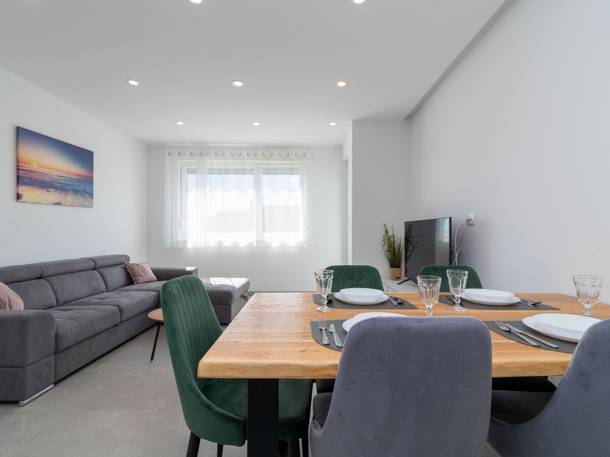 Ein moderner Essbereich mit einem Holztisch, gedeckt für zwei Personen, umgeben von grünen Polstersesseln. Ein graues Sofa steht an der Wand, und ein Flachbildfernseher ist rechts an der Wand befestigt.