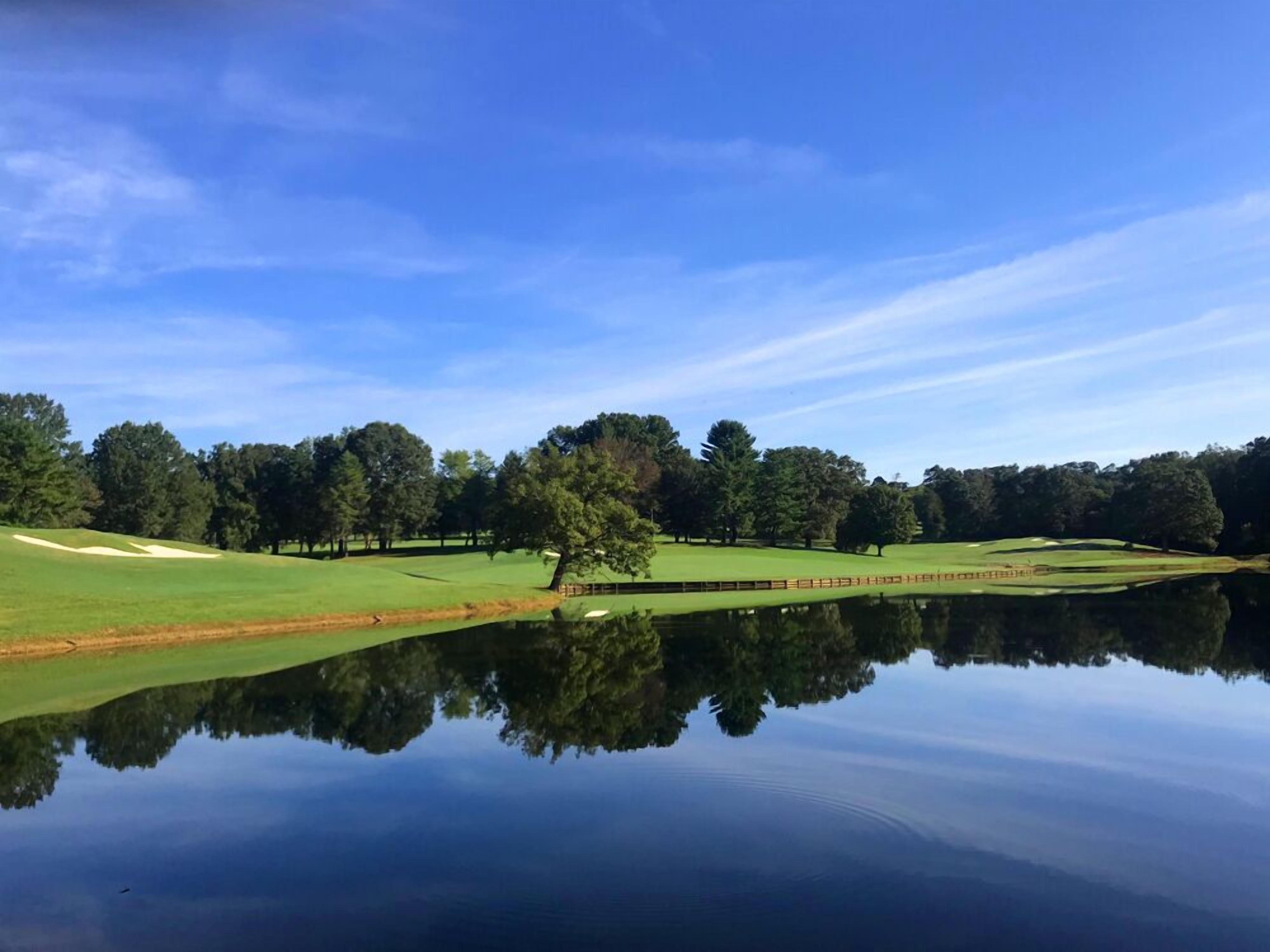 A serene golf course with a pond reflecting the landscape, surrounded by trees and a clear sky.