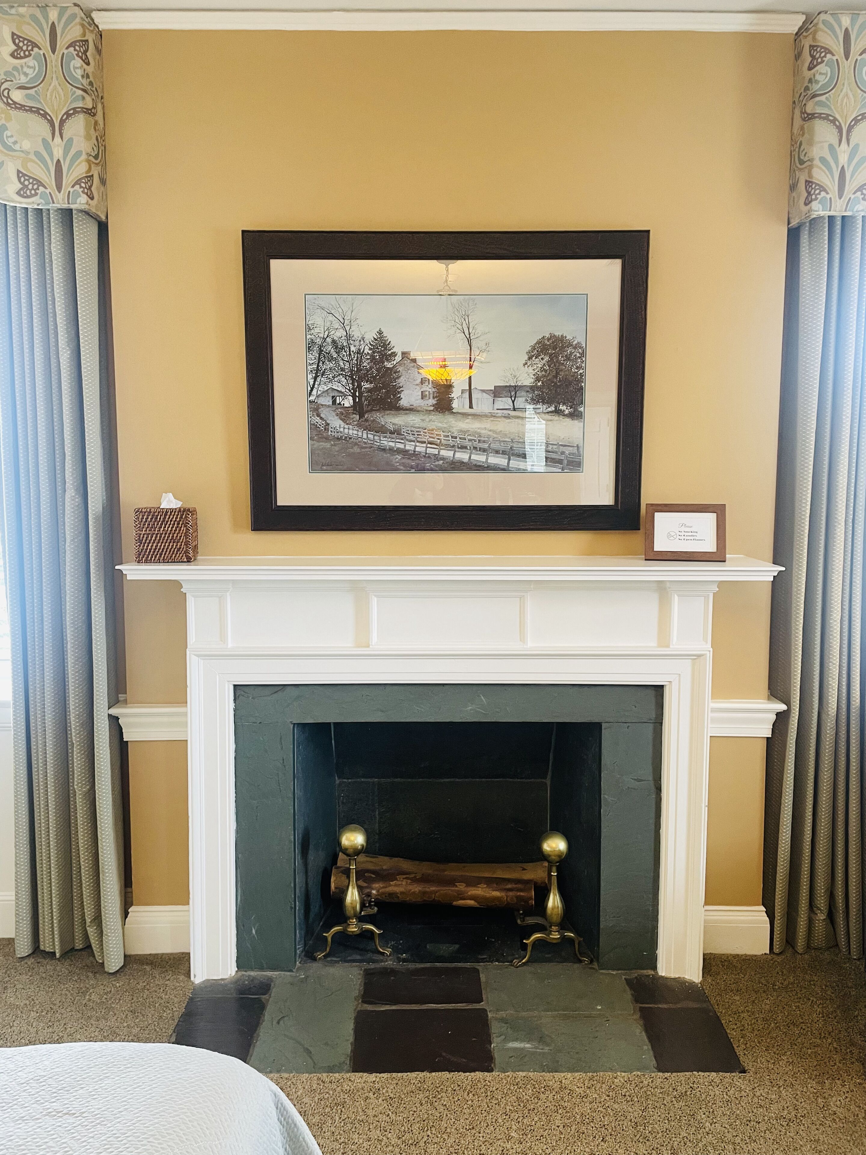 A fireplace with a framed picture above it, curtains on either side, and a small framed sign on the mantel.