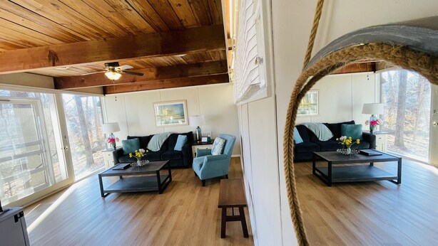 A spacious living room with wooden floors, a ceiling fan, and large windows offering a view of the outdoors. - Excelsior at the Manor House (Clemmons)