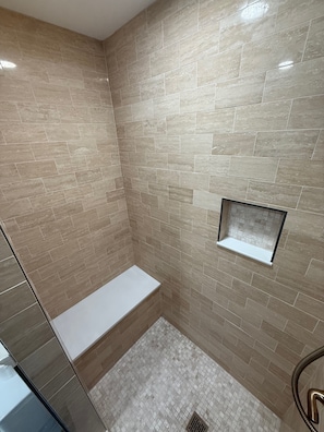 A bathroom with beige tiled walls, a white bench, and a small shelf.