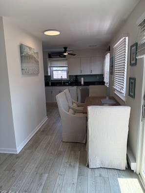 A modern kitchen with white cabinets, a dining area with two beige chairs, a ceiling fan, and a window with blinds.