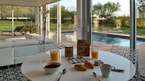 Comedor con una mesa dispuesta para el desayuno, incluyendo cruasanes, jugo y café, con vistas a una piscina y jardín.