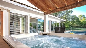 Zona de hidromasaje con terraza de madera, grandes puertas correderas de vidrio y vistas a una piscina y vegetación.