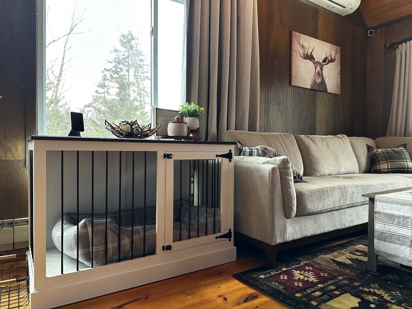 En hyggelig stue med en sofa, en tremmehegn til kæledyr, et vindue med gardiner og et billede af en hjort på væggen.