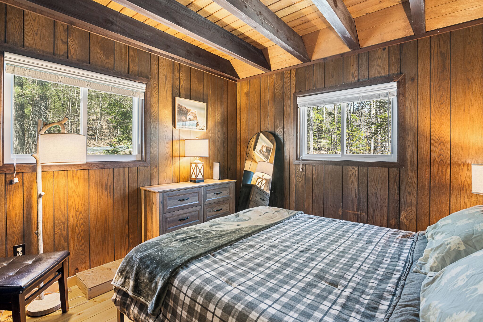 A bedroom with wooden walls, a bed with a plaid bedspread, a dresser, and two windows with views of trees.