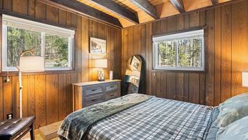 A bedroom with wooden walls, a bed with a plaid bedspread, a dresser, and two windows with views of trees.