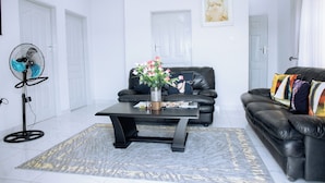 A modern living room with a black leather sofa, a black coffee table, and a standing fan. - Luxury 2 Bed Self Catering Apartment in Masvingo (Masvingo)