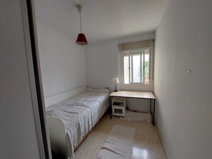 A small, sparsely furnished room with a single bed, a desk, and a window.