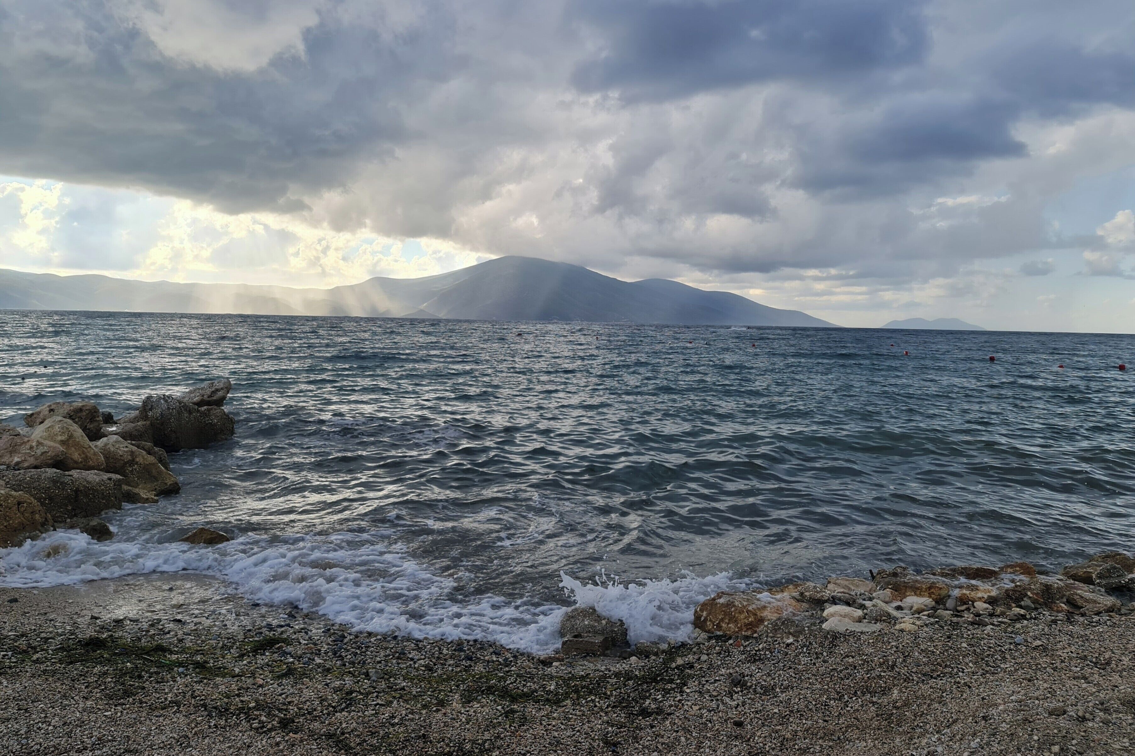 Un paysage côtier avec une rive rocheuse, une mer calme et des montagnes lointaines sous un ciel nuageux.
