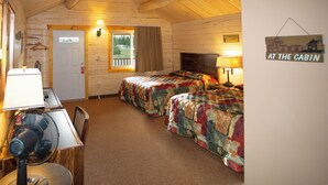 A cabin with two beds, a desk with a lamp, a fan, and a sign that reads "At the Cabin". - Young's Motel (Tok)