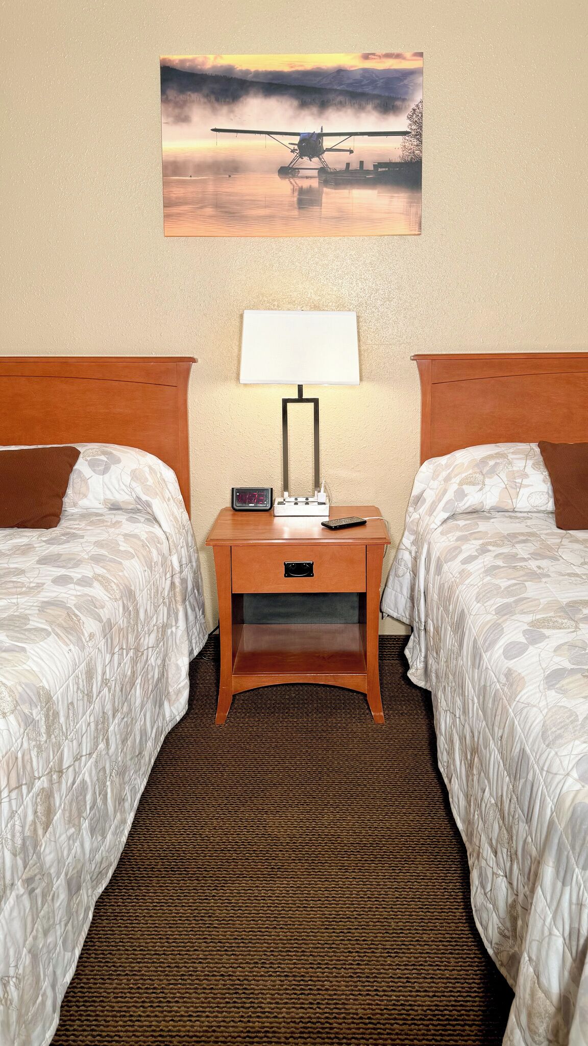A hotel room with two beds, a nightstand with a lamp, a clock, and a remote control.