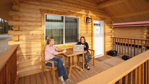 A wooden cabin with a deck, a table, and two people using a laptop. - Young's Motel (Tok)