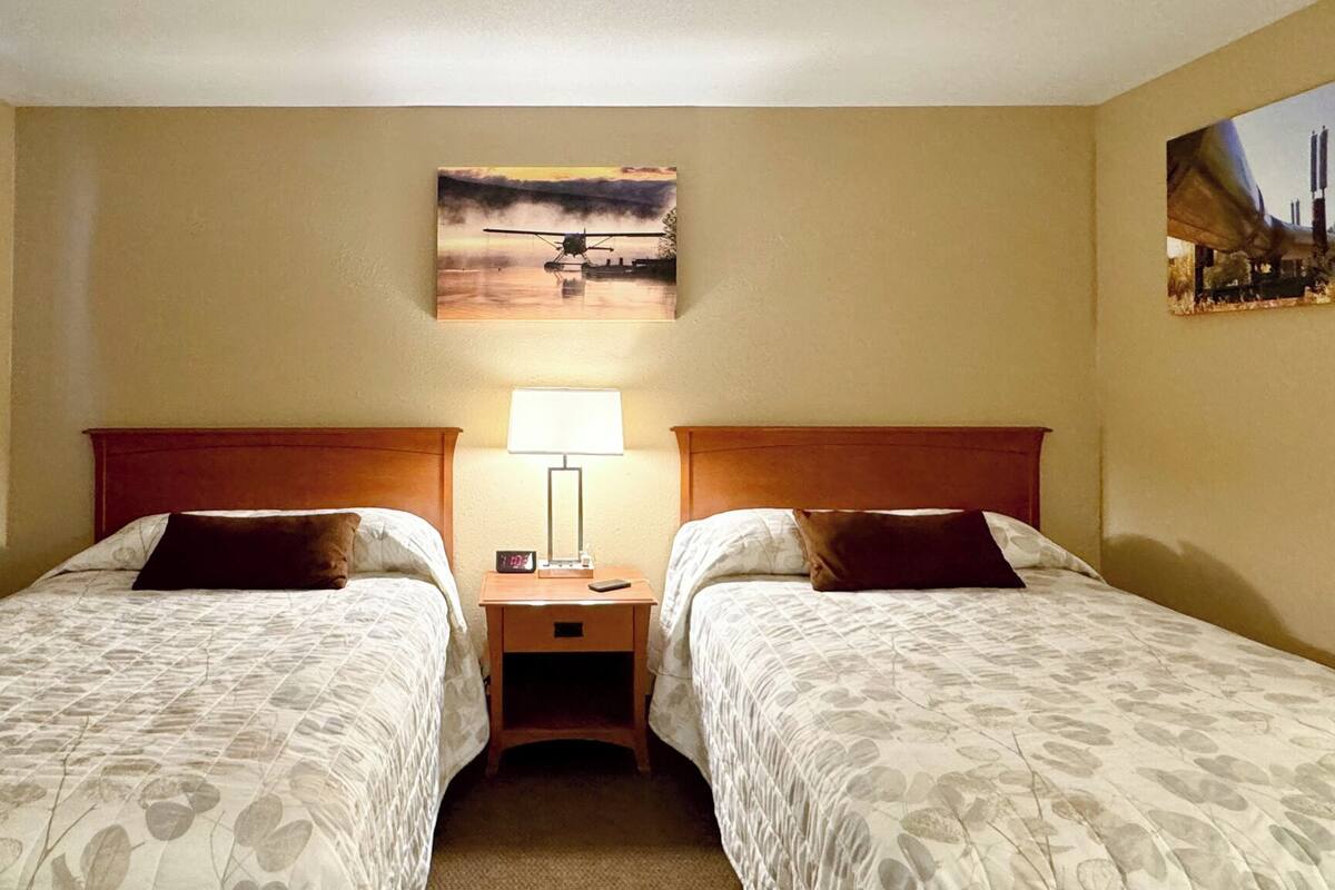 Quarto de hotel com duas camas, uma mesa de cabeceira com abajur e um quadro na parede.