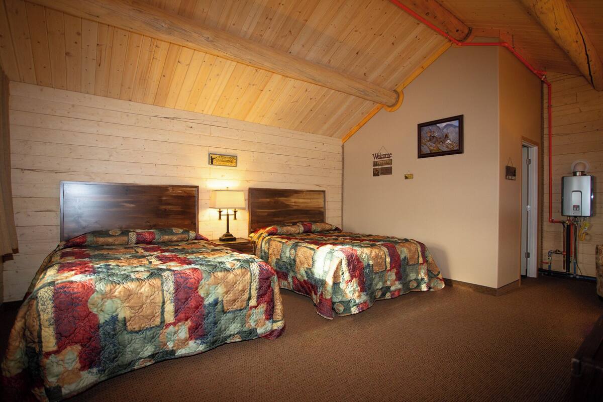 Quarto com duas camas, paredes de madeira, teto com vigas aparentes, placa de boas-vindas, quadro emoldurado e aquecedor de água.