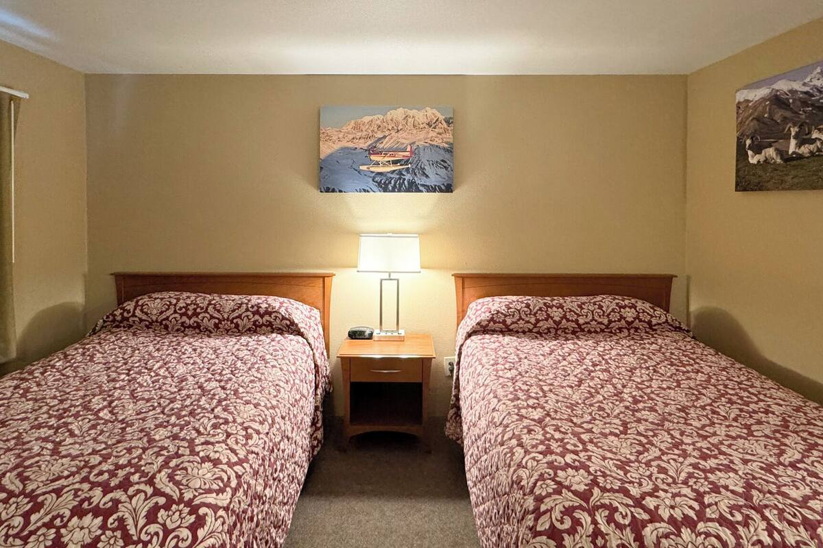Quarto de hotel com duas camas, uma mesa de cabeceira com abajur e um quadro de uma paisagem montanhosa na parede.