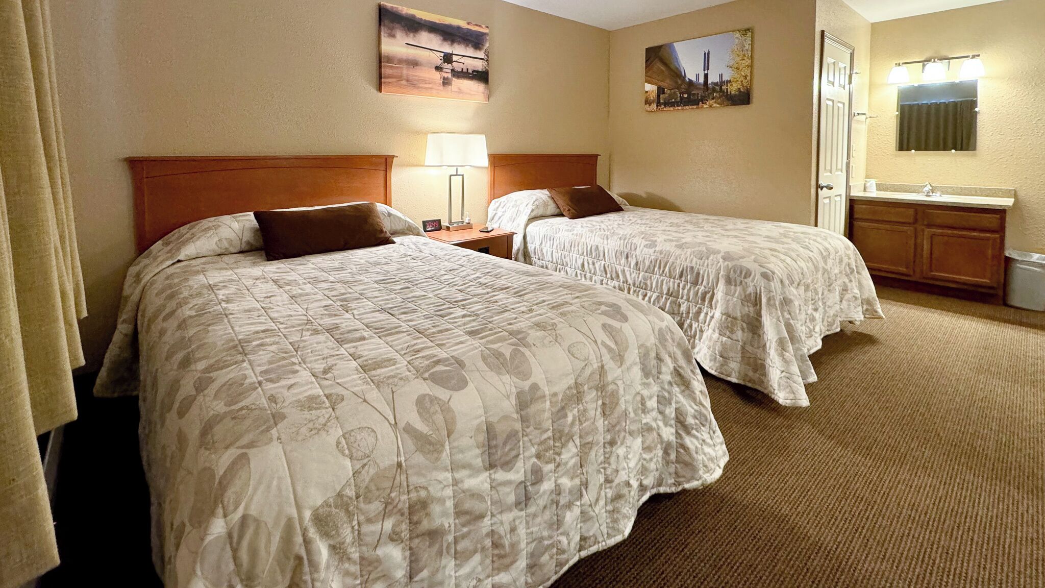 A hotel room with two beds, a nightstand, a lamp, a painting, a mirror, and a bathroom with a sink and vanity.
