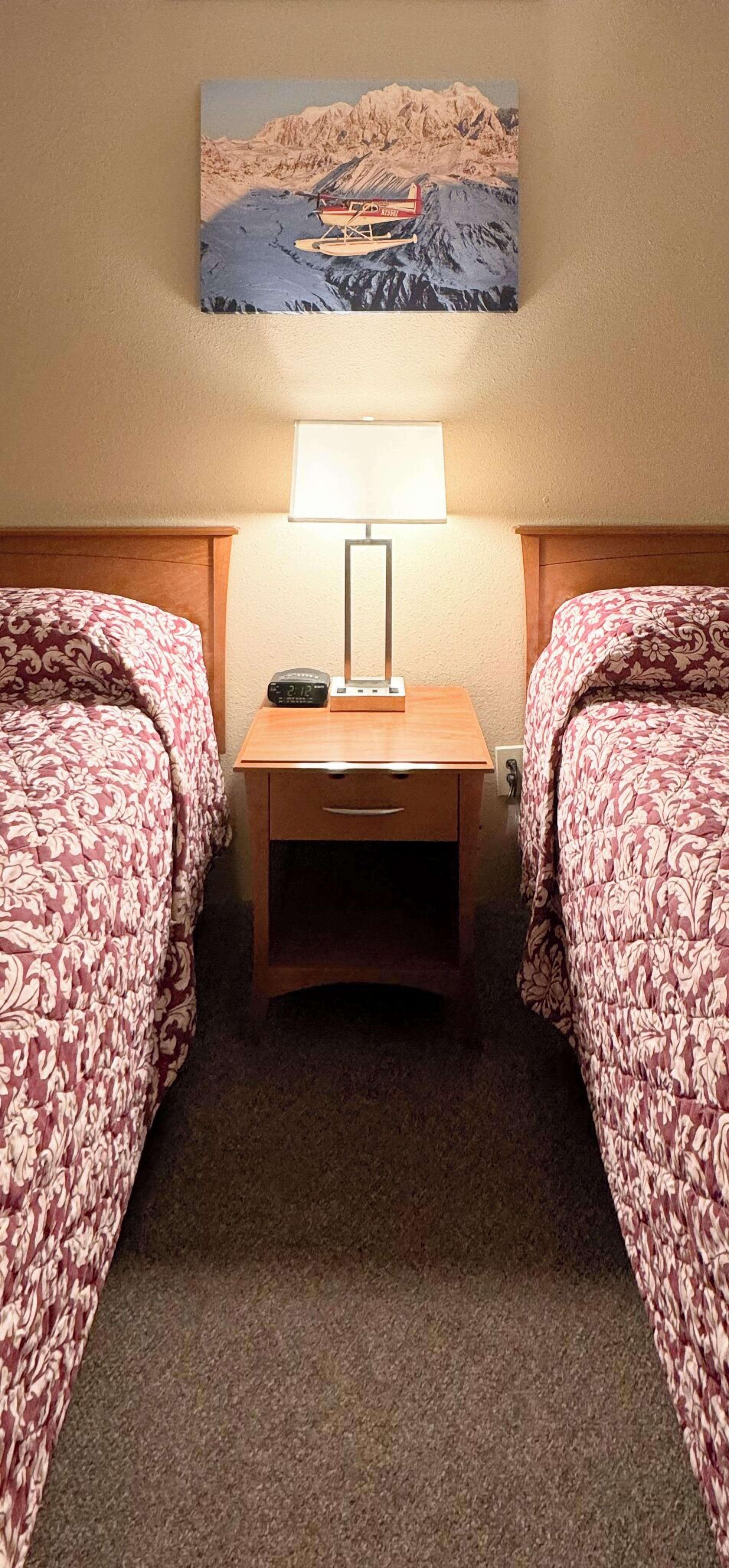 A hotel room with two beds, a nightstand with a lamp, and a painting of a mountainous landscape above the beds.