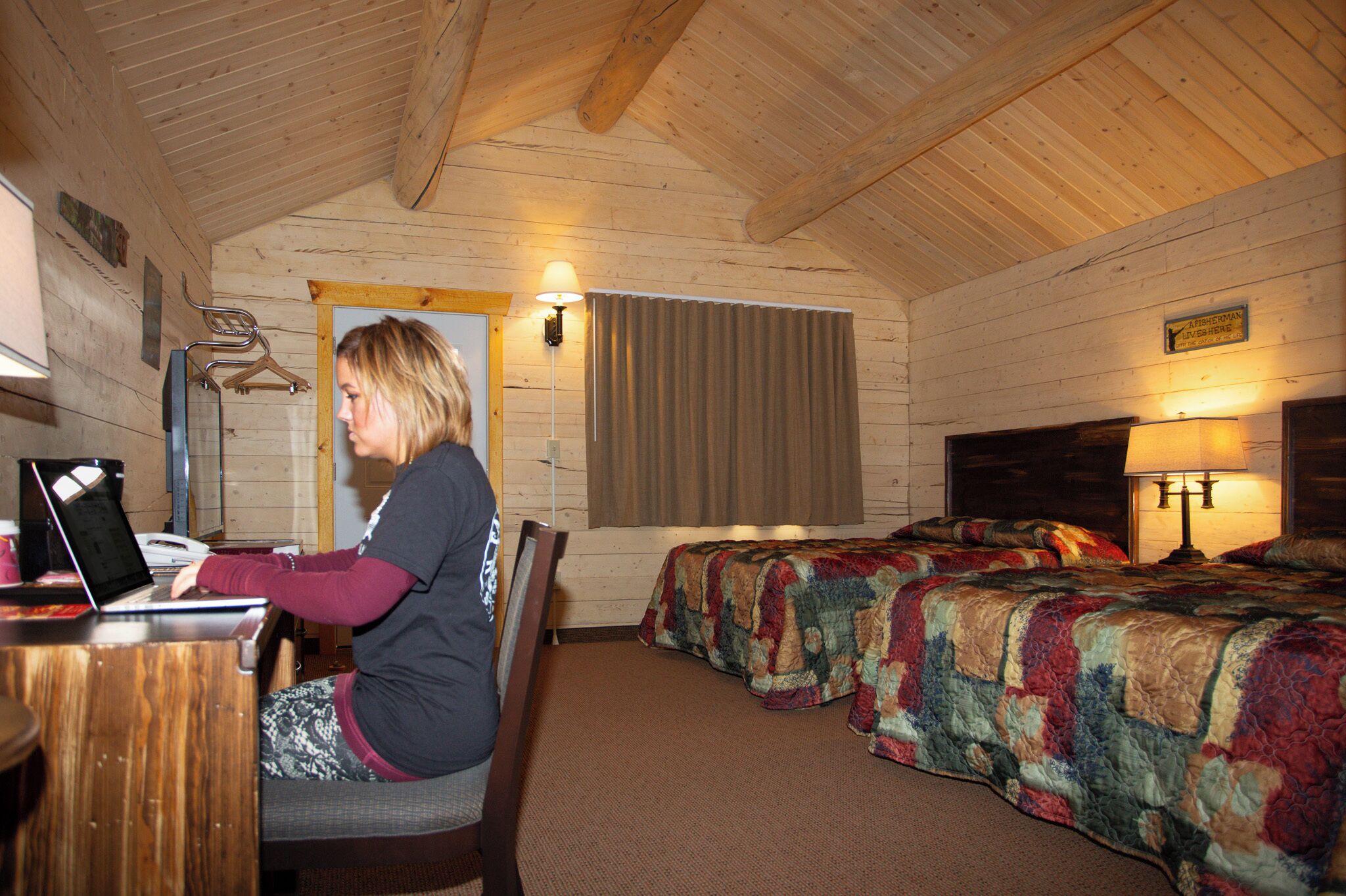 A room with wooden walls and a sloped ceiling, featuring two beds with colorful quilts, a desk with a laptop, and a lamp on the right side.
