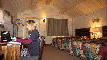 A room with wooden walls and a sloped ceiling, featuring two beds with colorful quilts, a desk with a laptop, and a lamp on the right side.